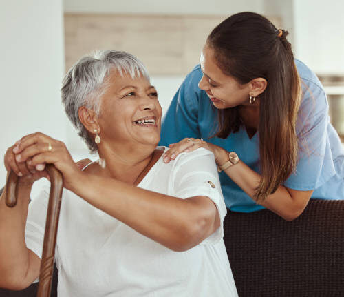 Happy, relax and senior woman with caregiver smile while sitting on a living room sofa in a nursing home. Support, help and professional nurse or healthcare worker helping elderly lady or patient